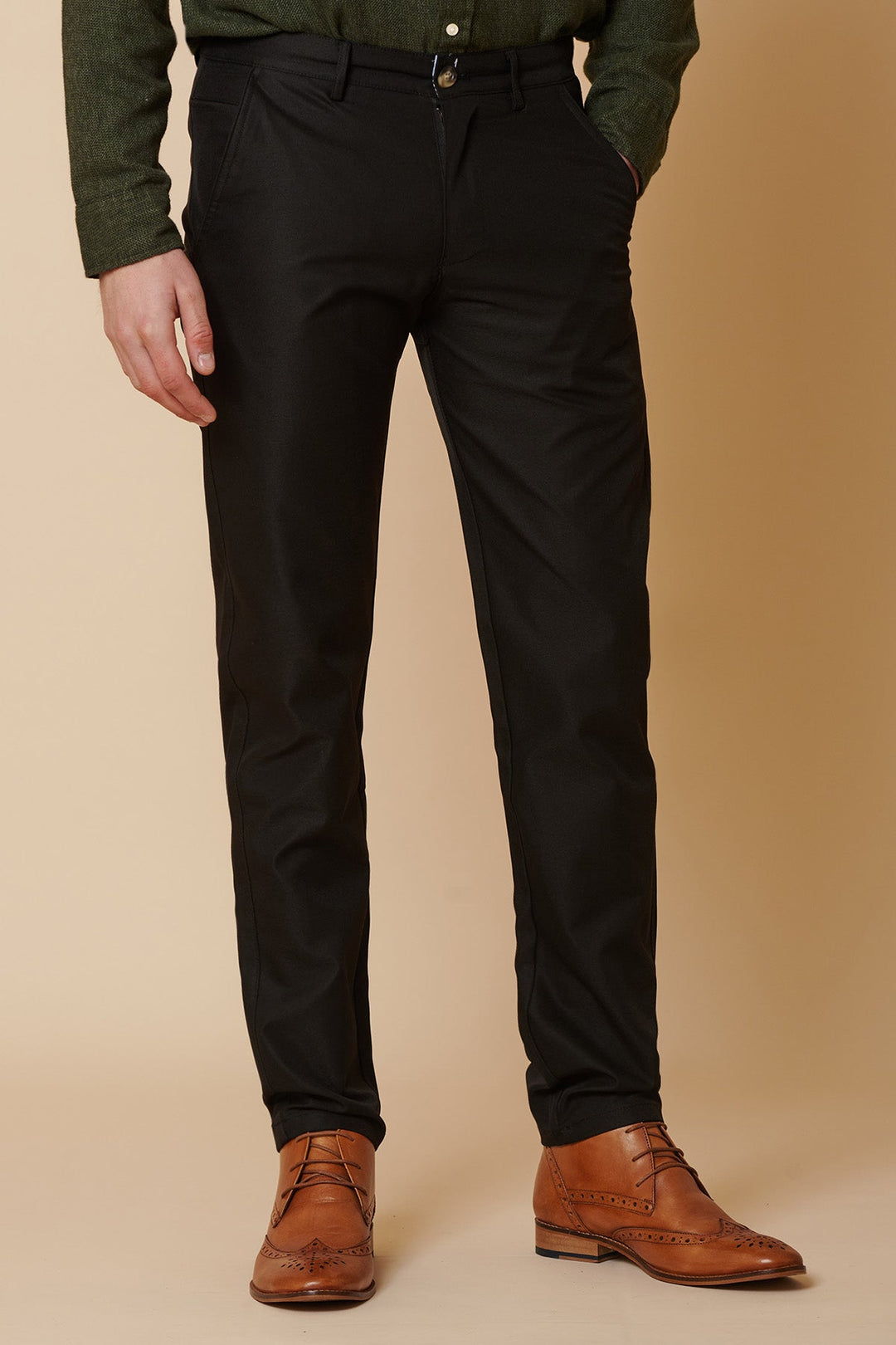 A person wearing black dress pants, a dark green shirt, and brown leather dress shoes stands with hands in pockets against a beige background. The image shows only the lower half of the body.