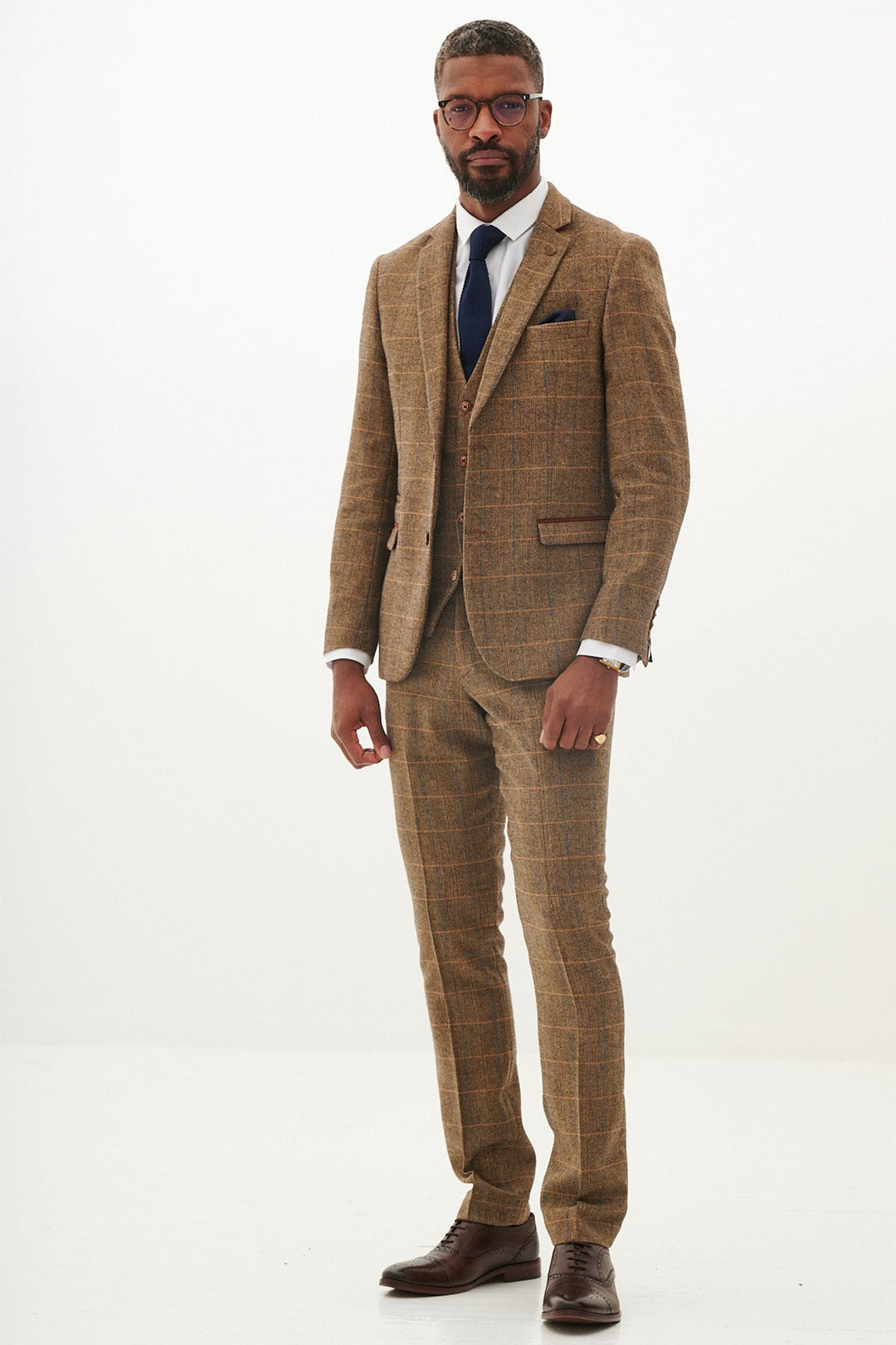 A man stands in a tan plaid three-piece suit with a white dress shirt, navy tie, brown shoes, and glasses against a plain white background.