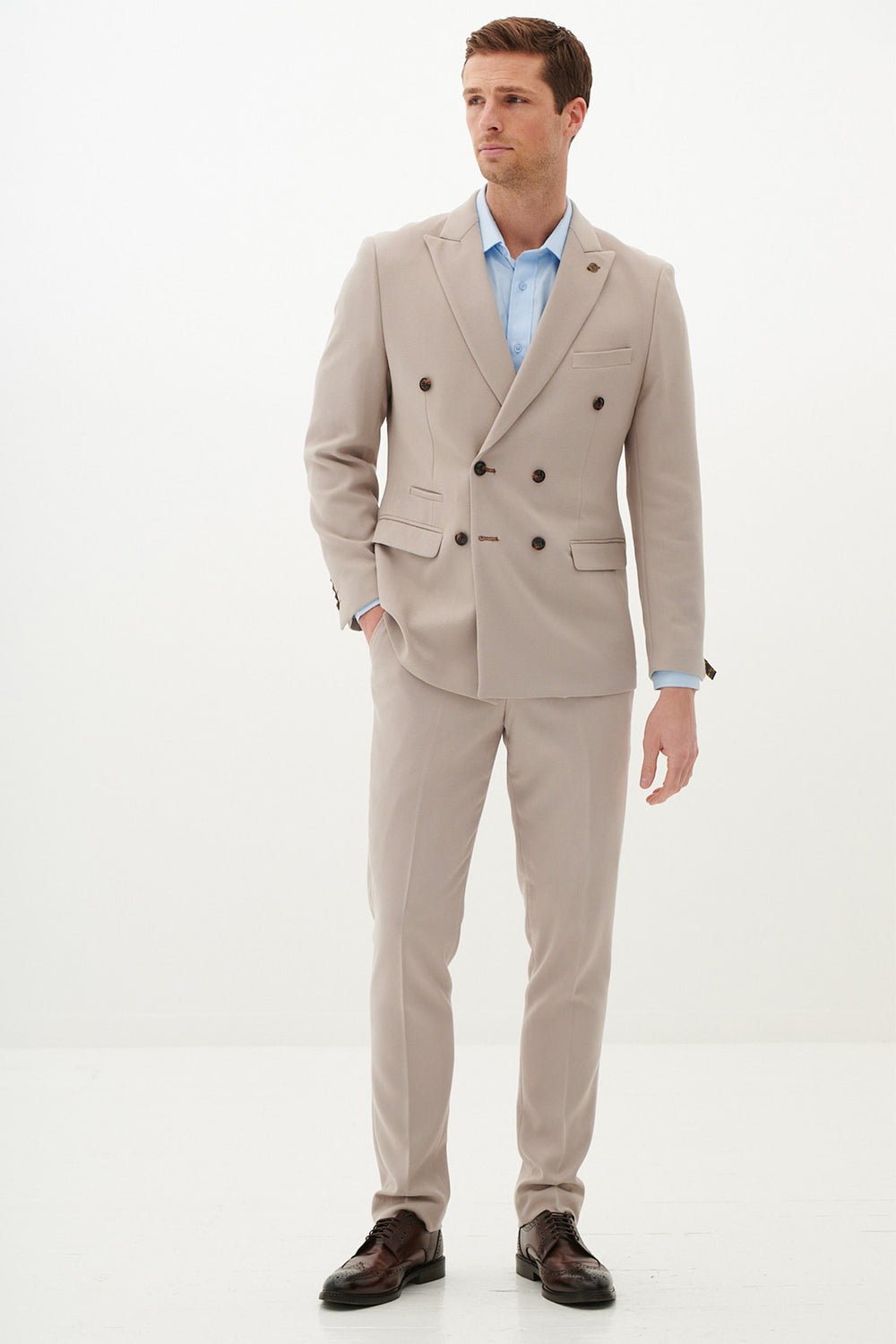 A man with short brown hair, looking slightly to the side, wears the Marc Darcy HM5 - Stone Double-Breasted Two Piece Suit in light beige poly-viscose, paired with a light blue shirt and brown dress shoes against a plain white background.