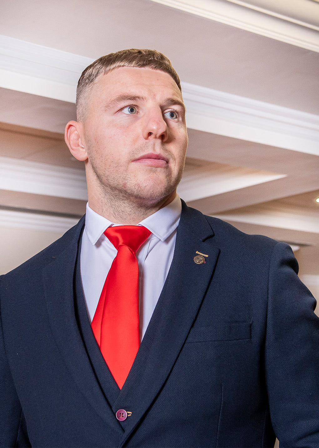 A man wearing a dark suit, white shirt, and bright red tie stands indoors, looking slightly to the side. The background shows a ceiling with recessed lighting and white trim.