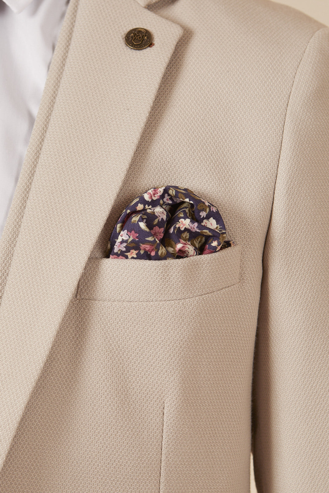 A close-up of the Marc Darcy HM5 - Stone Tailored Three Piece Suit features a slim fit jacket over a white shirt, accented by a floral pocket square in the breast pocket and a round lapel pin on the lapel.