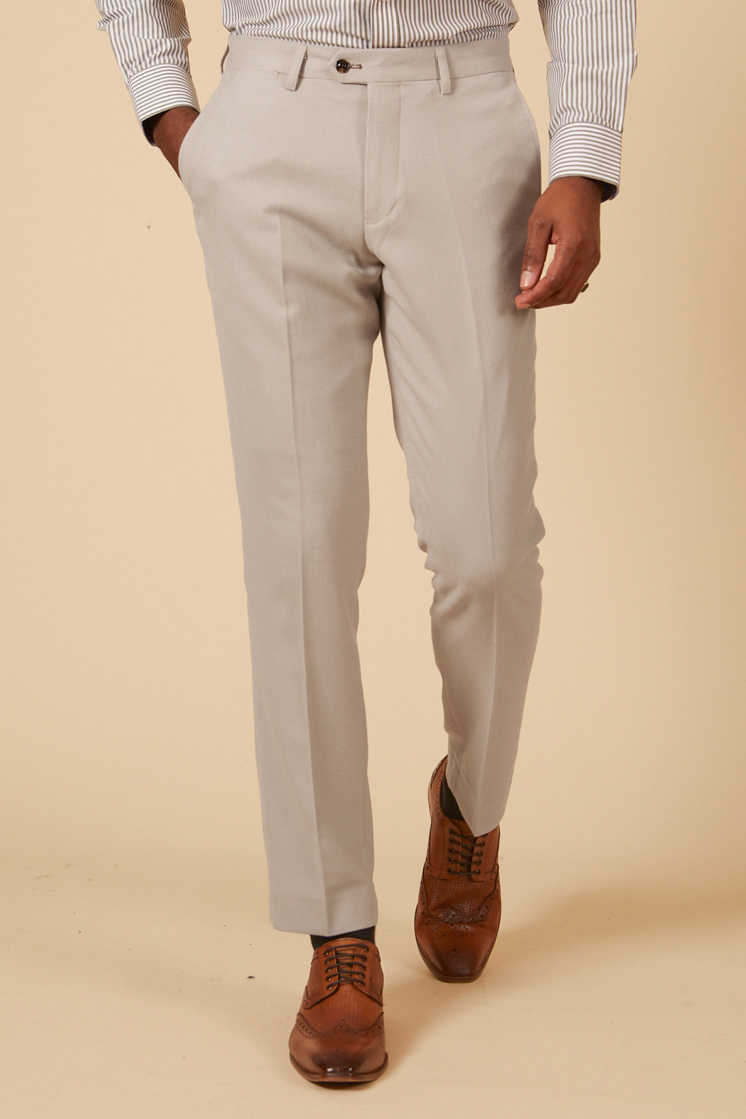 A person wearing Marc Darcys HM5 - Stone Tailored Three Piece Suit stands against a beige background. Only the lower half of the body is visible, showcasing slim fit suit pants, a striped shirt, and brown leather dress shoes.