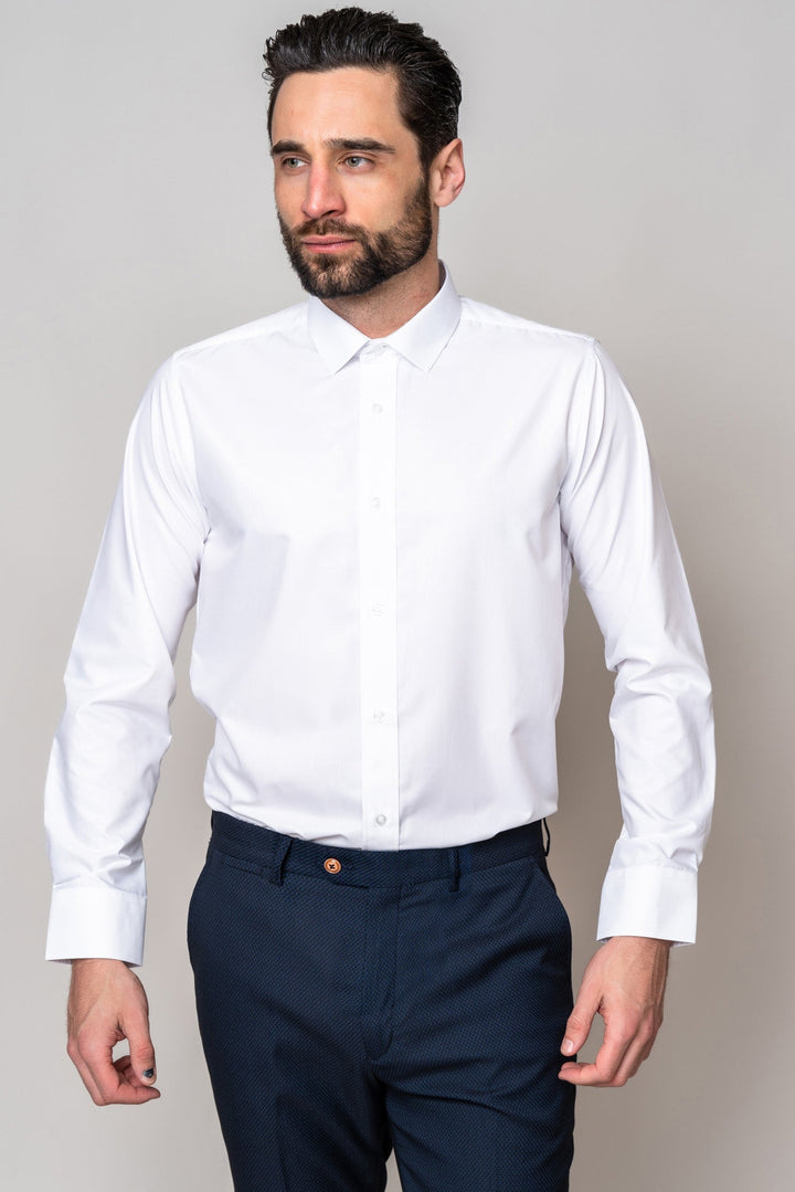 A man with dark hair and a beard wears the Marc Darcy CARTER White Long Sleeve Shirt with dark blue trousers. He stands against a light gray background, looking slightly to the side.